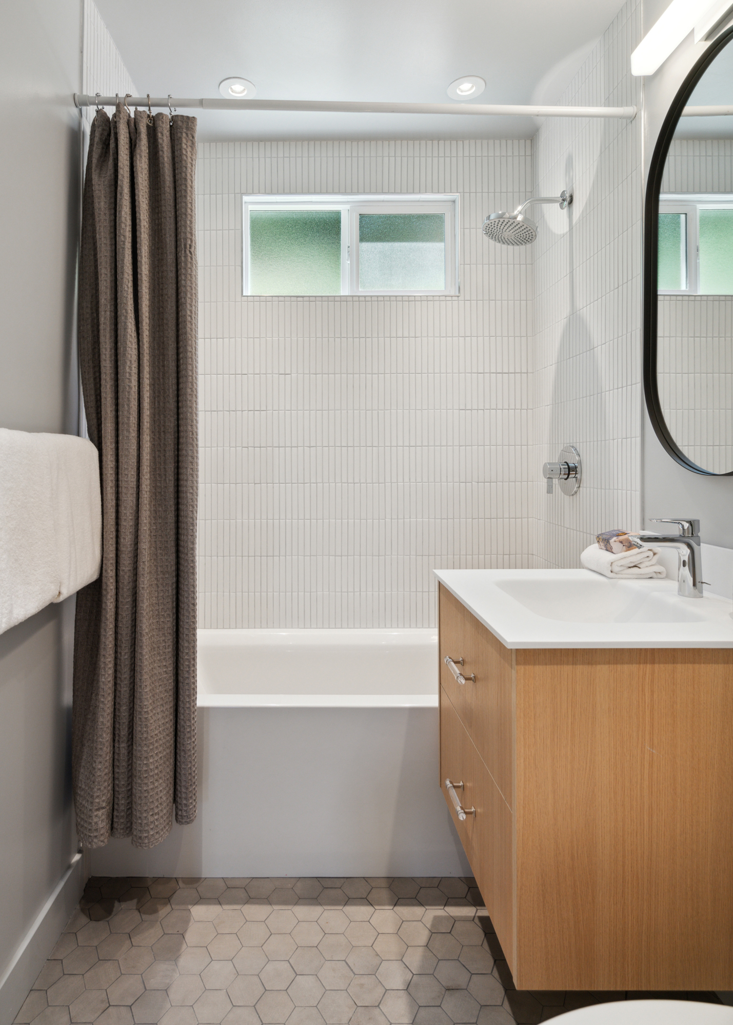 A three-piece bathroom with white tile, tub, floating oak vanity and hex tile floors in gray