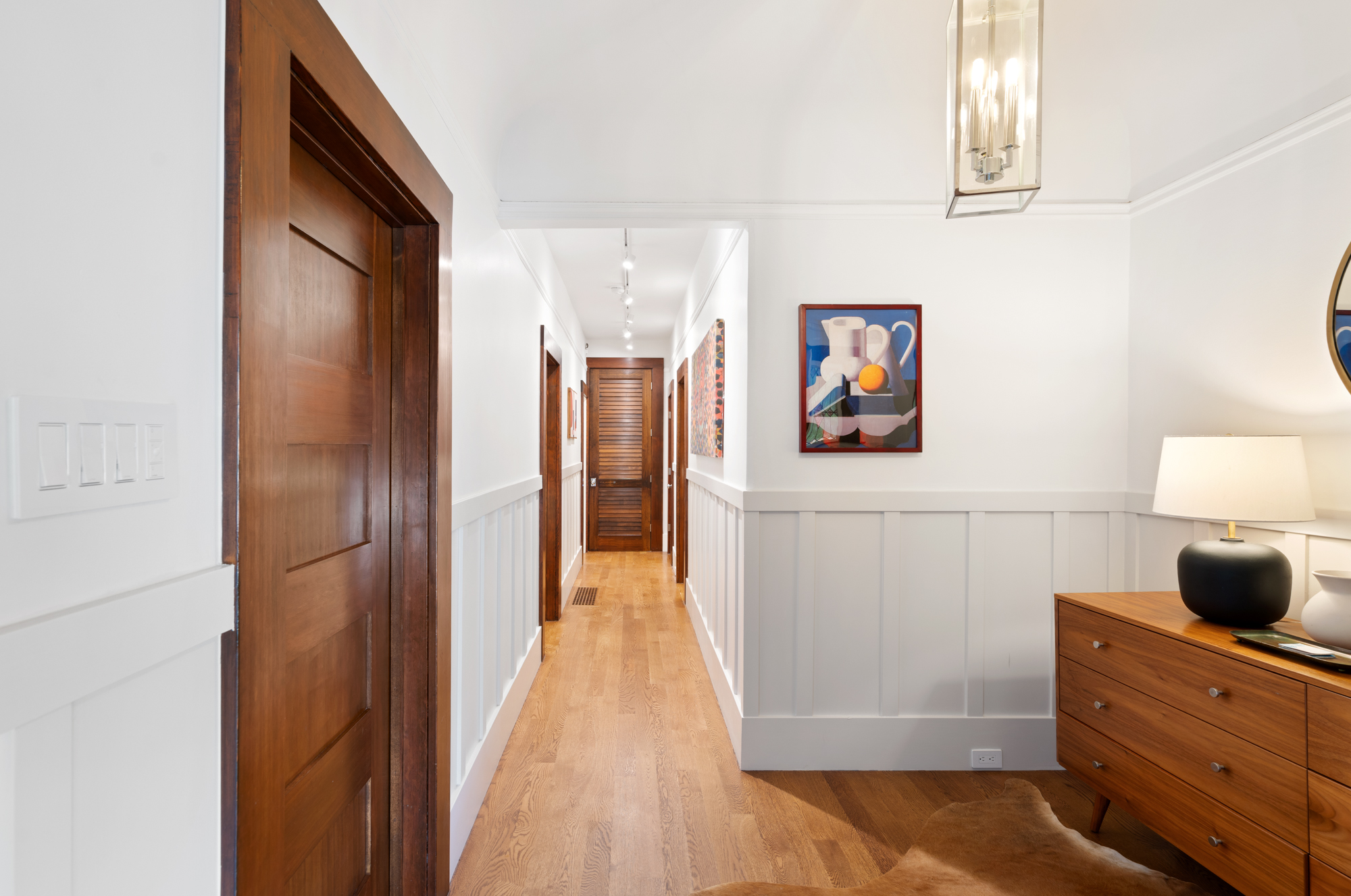 Long, welcoming hallway with hardwood floors, custom artwork, recessed lighting, and wood-trimmed doors