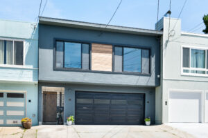 Street-facing exterior of a two-level home with dark grey stucco, black trim, wood accent paneling, and a double garage.